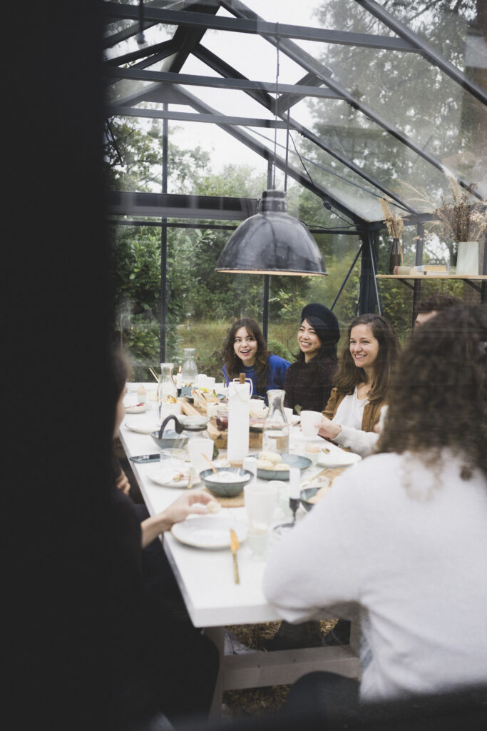 Gruppe von lachenden Frauen bei einem gemütlichen Essen in einem lichtdurchfluteten Glashaus, umgeben von Natur und Grün - Bahnhofszeit Foto: Maria Bayer https://www.mariabayer.de