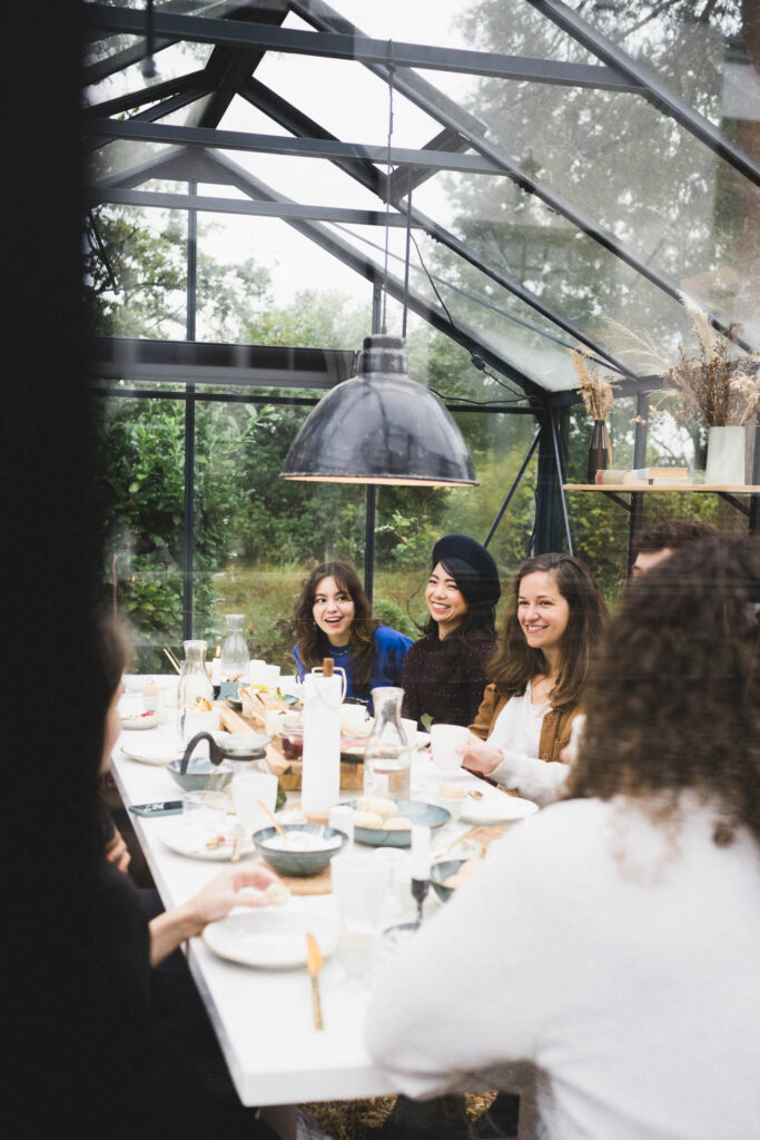 Gruppe von lachenden Frauen bei einem gemütlichen Essen in einem lichtdurchfluteten Glashaus, umgeben von Natur und Grün.