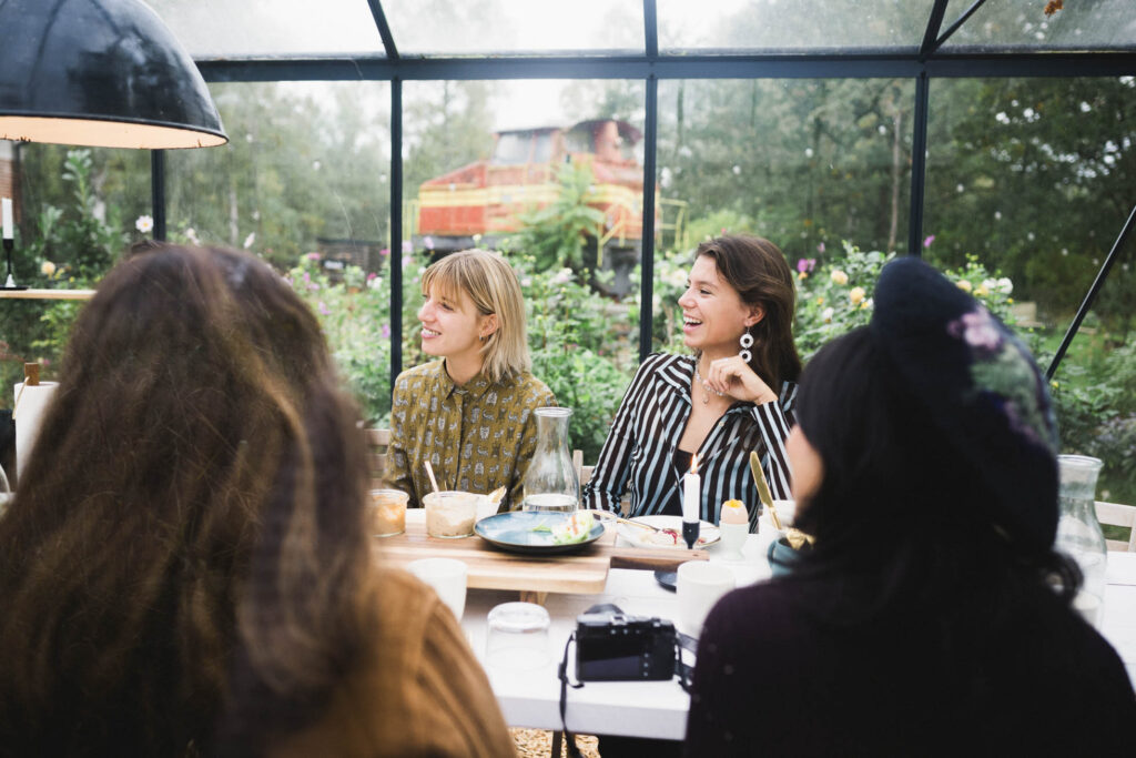 Fröhliche Teilnehmerinnen beim Frühstück im Glashaus von Bahnhofszeit, mit Blick auf Garten und historische Lok. Foto: Maria Bayer https://www.mariabayer.de