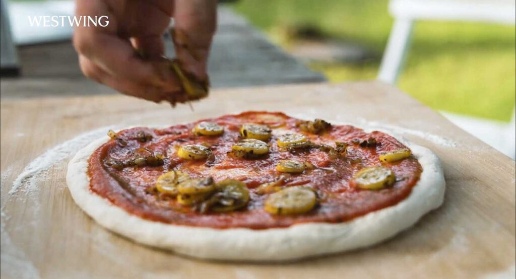Hand belegt eine frische Pizza mit Tomatensauce und Zucchini auf Holzbrett im Garten während eines Westwing Outdoor-Cooking-Shootings.