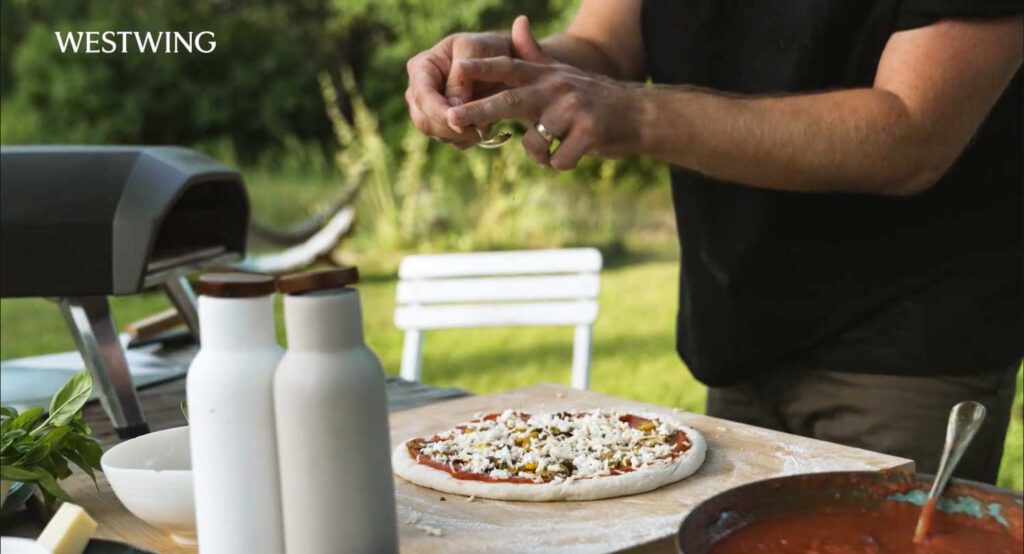 Hände streuen Zutaten auf eine Pizza im Garten, flankiert von Flaschen, Kräutern und einem Ooni-Ofen im Westwing Video.