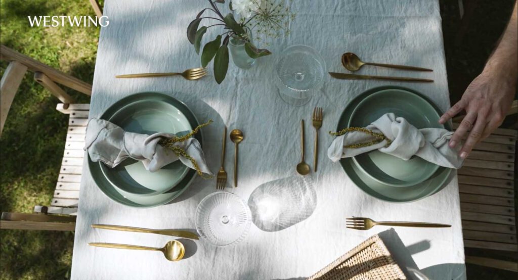 Draufsicht auf sommerlich gedeckten Tisch mit grünem Geschirr, goldenen Besteck und weißen Leinenservietten im Westwing Video.